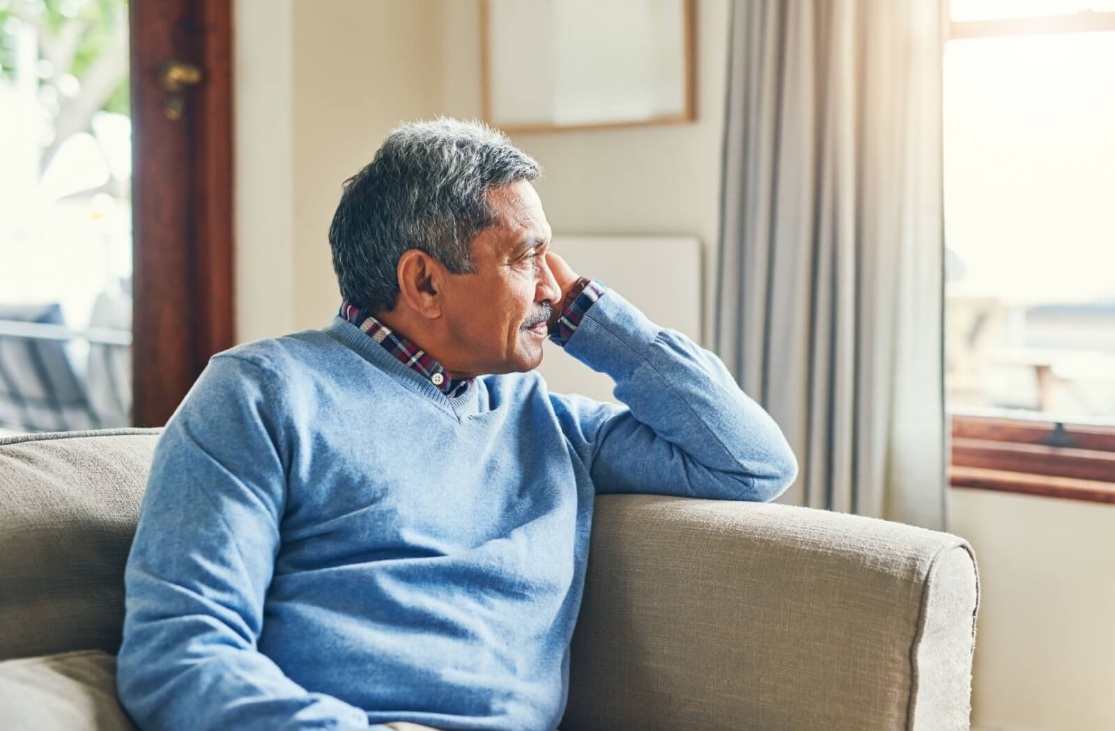 A senior with dementia sits at home alone, looking out a nearby window.