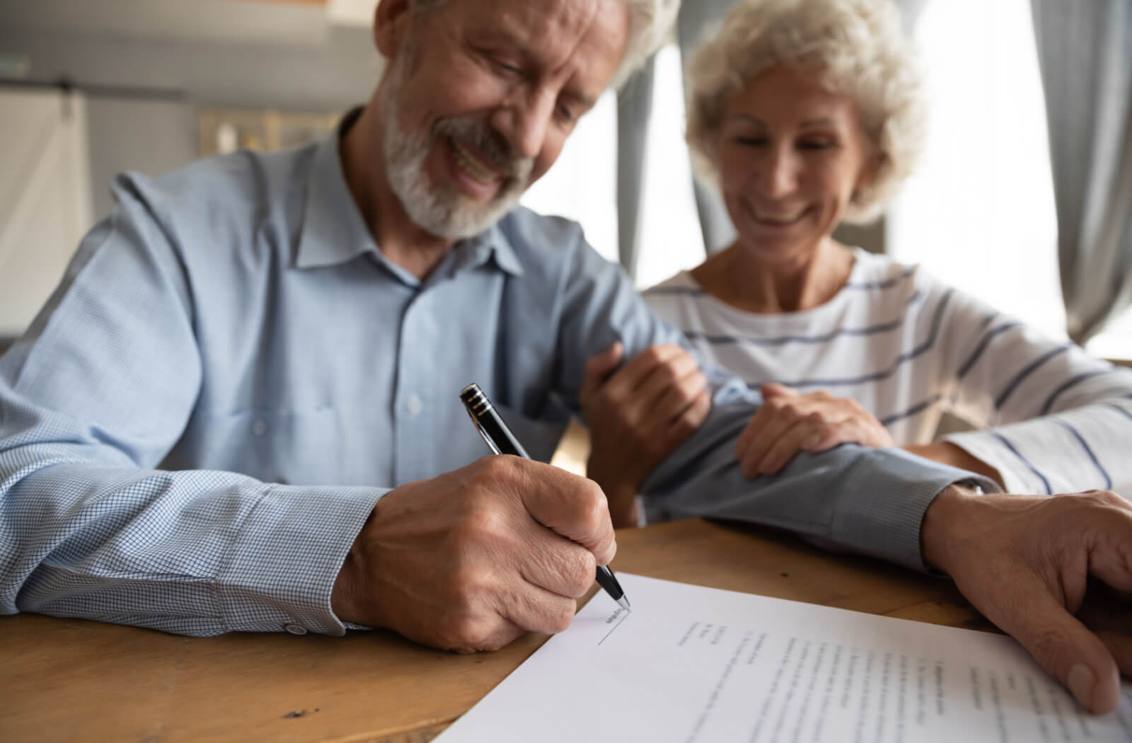 An older adult man signing a document while his wife happily holds his arm.