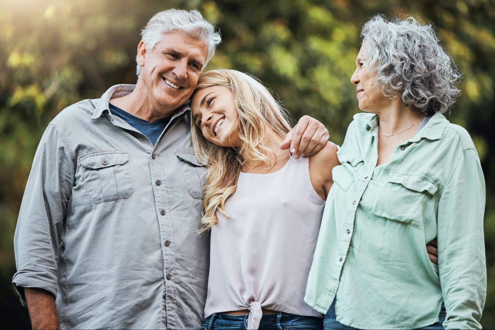 Two senior parents walking along an outdoor pathway with their daughter. The father has his arm over the daughter's shoulders, and all are smiling at the bright, sunny weather.