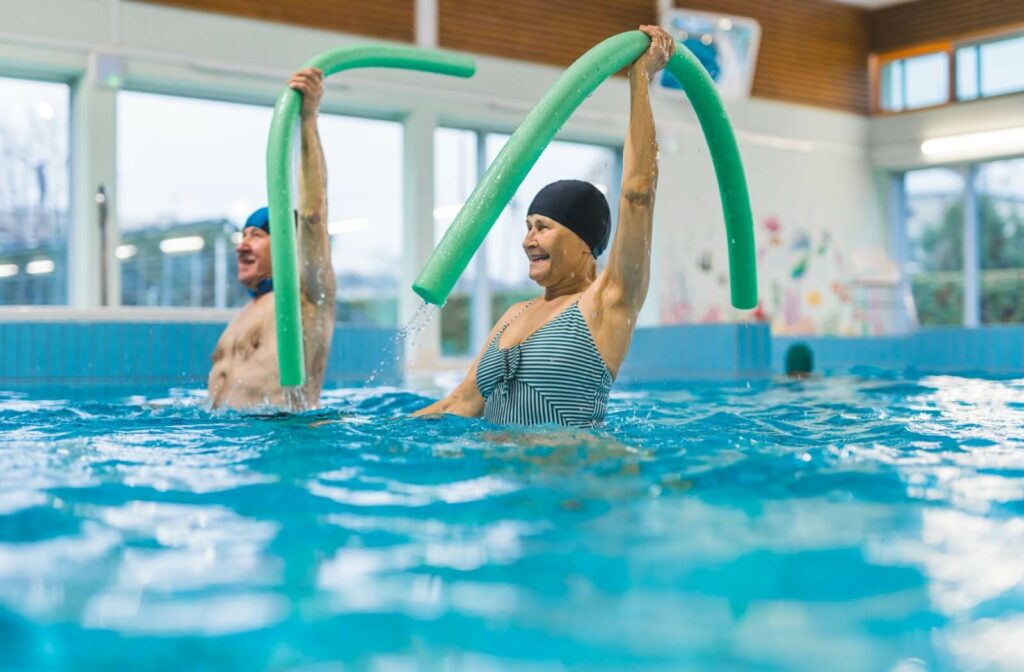 Two seniors in swimming caps in an indoor pool hold up green pool noodles as part of a water aerobics class