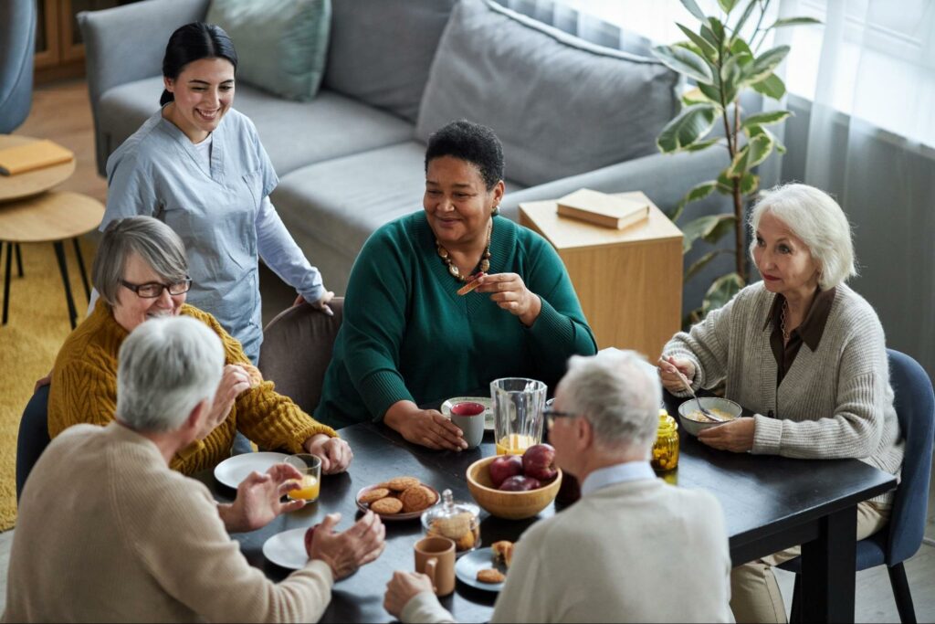 A group of seniors smiling and chatting while they eat breakfast in a well-lit dining room. A staff member is standing nearby with a welcoming smile.