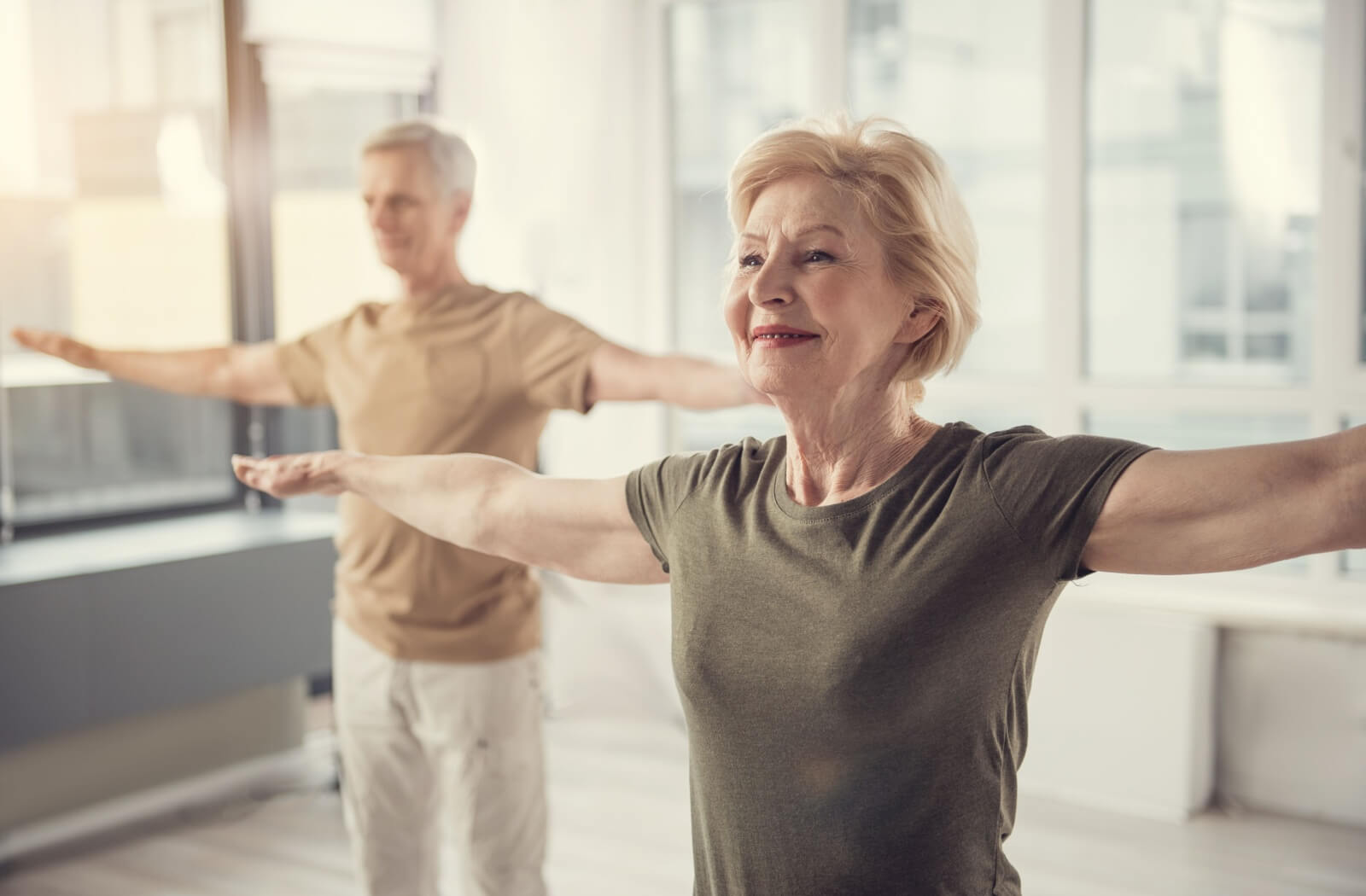 A senior couple stretch their arms out in a yoga pose to stretch in a cleared out indoor space