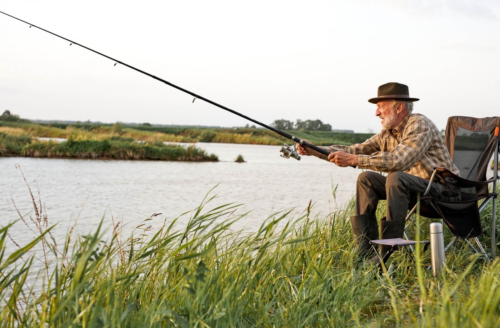 A senior sits by a lake in the early morning and casts their fishing line into the water.