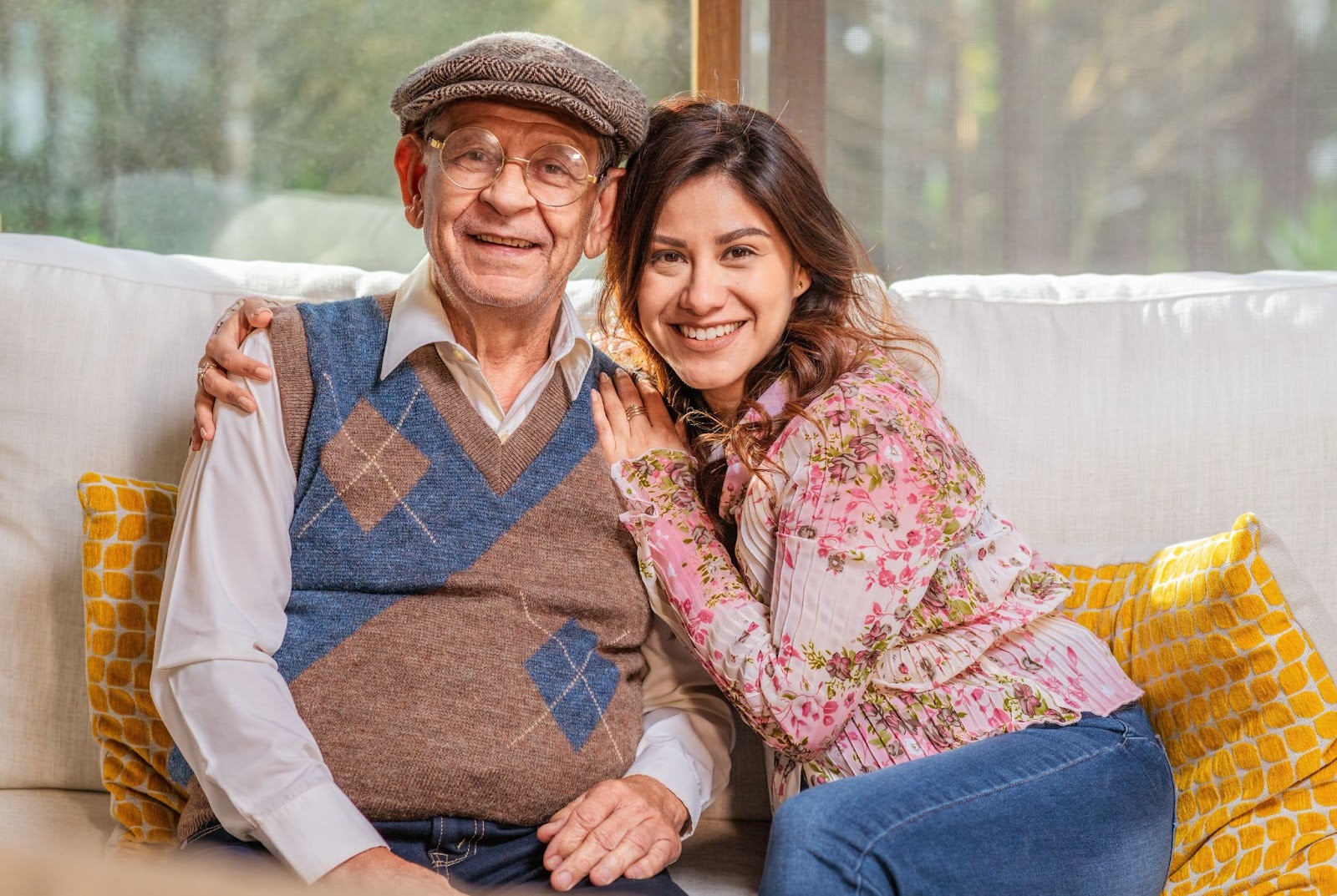 A young adult caregiver sits on the couch with an older adult and wraps their arms around the older adult's shoulders.