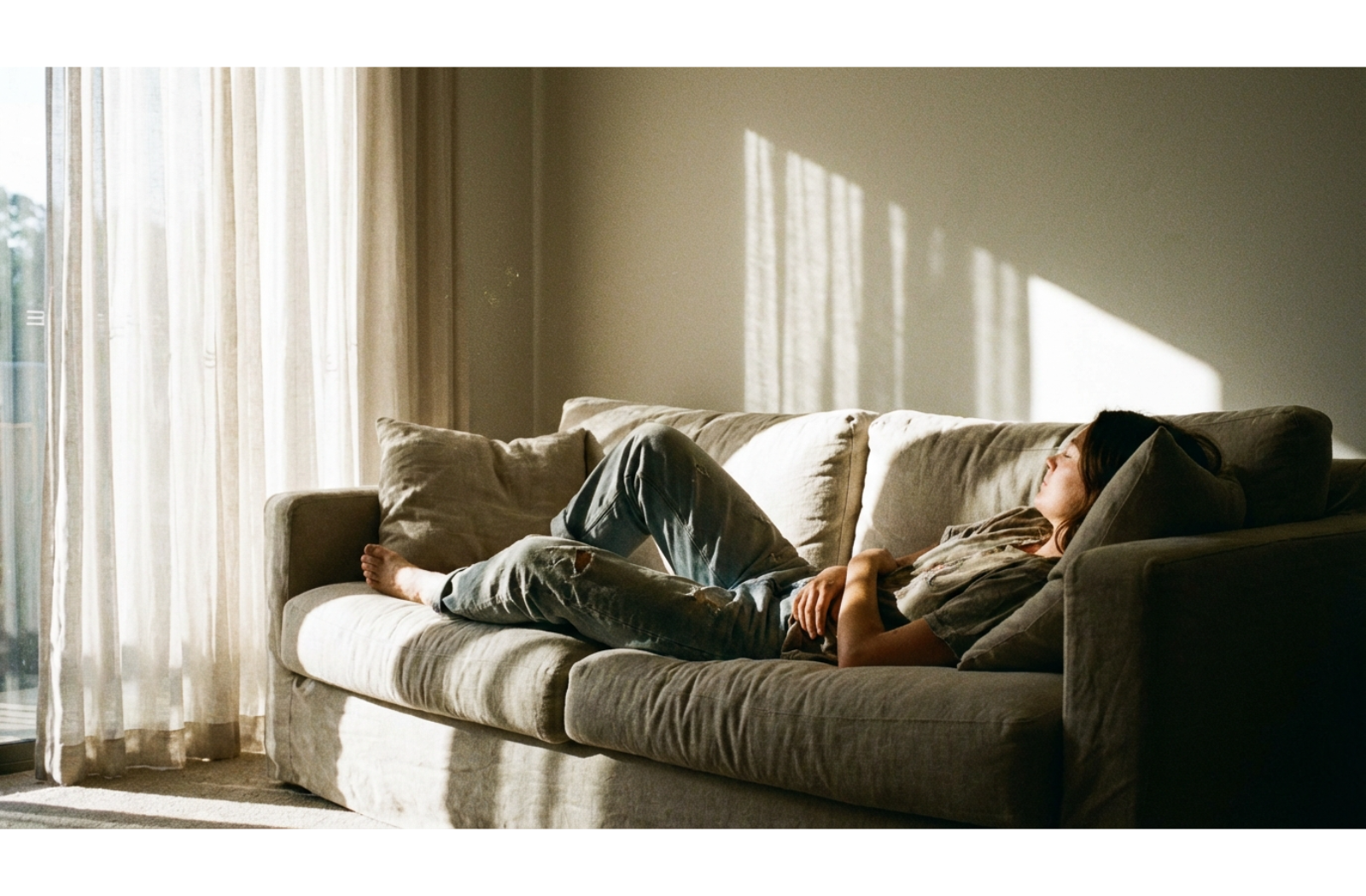 A person lounges on a linen sofa in a sunlit room, bathed in natural light and shadows during a quiet moment of relaxation.